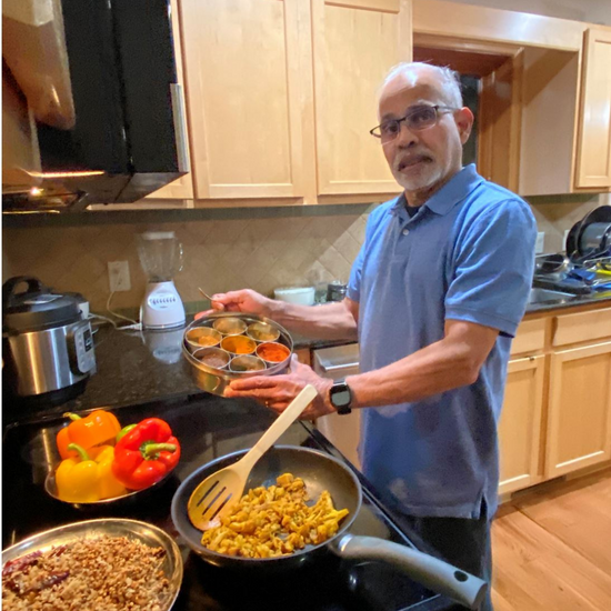 Picture of Dad cooking Indian Food with few spices and vegetables on the kitchen counter. Dad has  spice box his hand and looks confused at the number of spices in the box